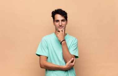 Genç yakışıklı adam mutlu görünüyor ve gülümsüyor. Elleri çenesinde, merak ediyor ya da soru soruyor. Seçenekleri karşılaştırıyor.