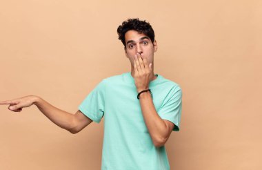 Genç yakışıklı adam kendini mutlu, şaşkın ve şaşırmış hissediyor. Ağzını eliyle kapatıyor ve yanal kopya alanını işaret ediyor.