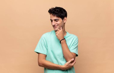 Genç ve yakışıklı bir adam mutlu, kendinden emin bir ifadeyle gülümsüyor. Elleri çenesinde, merak ediyor ve yan tarafa bakıyor.