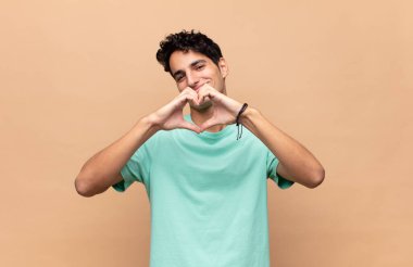 Genç yakışıklı adam gülümsüyor ve mutlu, sevimli, romantik ve aşık, iki eliyle kalbi şekillendiriyor.