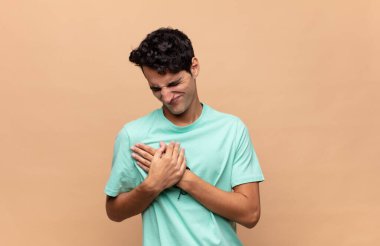 Genç yakışıklı adam üzgün, yaralı ve kalbi kırık görünüyor, iki elini de kalbine yakın tutuyor, ağlıyor ve bunalımda.