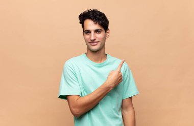 Genç yakışıklı adam neşeyle gülümsüyor, mutlu hissediyor ve kenarı ve yukarıyı işaret ediyor, fotokopi uzayındaki nesneleri gösteriyor.