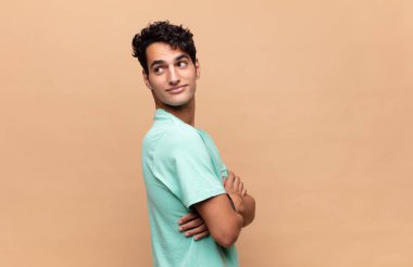 Genç yakışıklı adam neşeyle gülümsüyor, mutlu, tatmin olmuş ve rahatlamış hissediyor, kollarını kavuşturup yan tarafa bakıyor.