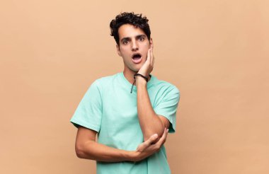 Genç yakışıklı adam şok ve inançsızlık içinde, elleri yanak ve kolları çapraz, sersemlemiş ve şaşırmış hissediyor.