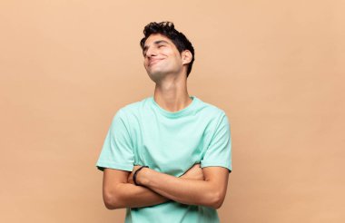 Genç yakışıklı adam kollarını kavuşturup mutlu bir şekilde gülüyor, rahat, pozitif ve tatmin edici bir pozla.