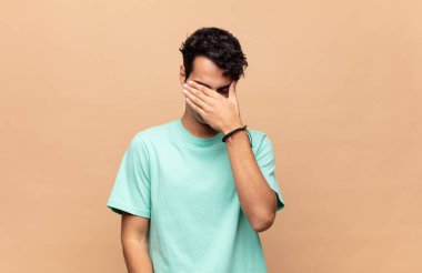 Genç ve yakışıklı bir adam stresli, utanmış ya da üzgün görünüyor, baş ağrısı var, yüzünü eliyle kapatıyor.