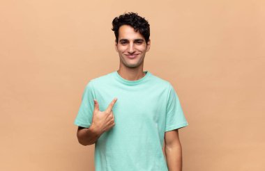 Genç yakışıklı, gururlu, kendinden emin ve mutlu görünüyor, gülümsüyor ve kendini işaret ediyor ya da bir numara işareti yapıyor.