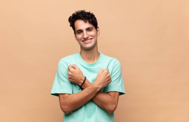 Genç yakışıklı adam, neşeli bir şekilde gülümsüyor ve kutluyor. Yumruklarını sıkmış, kollarını kavuşturmuş, mutlu ve pozitif hissediyor.