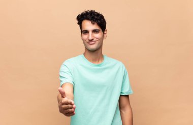 Genç yakışıklı adam gülümsüyor, mutlu görünüyor, kendinden emin ve arkadaş canlısı, bir anlaşmaya varmak için el sıkışıyor, işbirliği yapıyor.