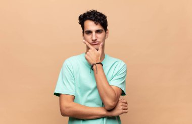 Genç yakışıklı adam ciddi, düşünceli ve güvensiz görünüyor, bir kolu çapraz ve çene üzerinde, seçenekleri tartıyor