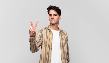 Genç yakışıklı adam gülümsüyor ve mutlu görünüyor, kaygısız ve olumlu, zafer veya barış işareti yapıyor.