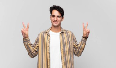 Genç yakışıklı adam gülümsüyor ve mutlu görünüyor, arkadaş canlısı ve memnun, işaret eden zafer ya da barış iki elle