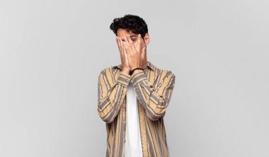 Genç yakışıklı adam elleriyle yüzünü kapatıyor, parmaklarının arasından şaşkın bir ifadeyle yan tarafa bakıyor.