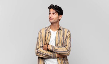 Genç yakışıklı adam omuz silkiyor, kafası karışık ve kararsız, kolları bağlı ve şaşkın görünüyor.