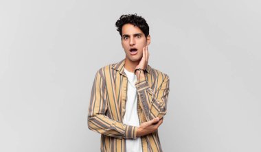 Genç yakışıklı adam şok ve inançsızlık içinde, elleri yanak ve kolları çapraz, sersemlemiş ve şaşırmış hissediyor.