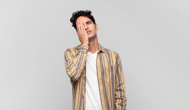 Genç yakışıklı adam uykulu, sıkılmış ve esniyor, baş ağrısı ve yüzünün yarısını kaplayan bir elle.