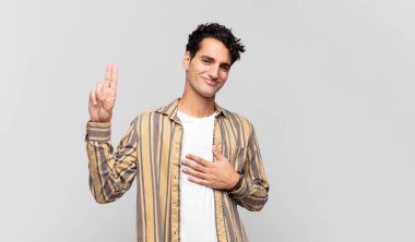 Genç yakışıklı adam mutlu, kendinden emin ve güvenilir görünüyor, gülümsüyor ve zafer işareti gösteriyor, olumlu bir tavırla.