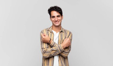 Genç yakışıklı adam, neşeli bir şekilde gülümsüyor ve kutluyor. Yumruklarını sıkmış, kollarını kavuşturmuş, mutlu ve pozitif hissediyor.