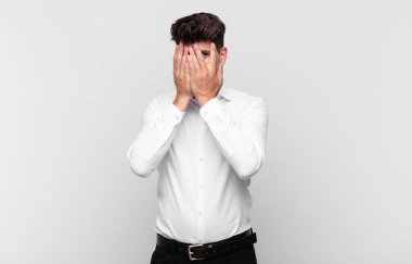 Genç yakışıklı adam elleriyle yüzünü kapatıyor, parmaklarının arasından şaşkın bir ifadeyle yan tarafa bakıyor.
