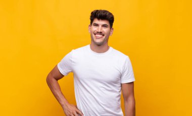 Genç yakışıklı adam mutlu bir şekilde gülümsüyor, eli kalçasında ve kendinden emin, olumlu, gururlu ve arkadaş canlısı.