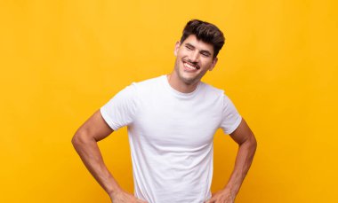 Genç yakışıklı adam mutlu, neşeli ve kendinden emin görünüyor. Gururla gülümsüyor ve iki elini de kalçasında istiyor.