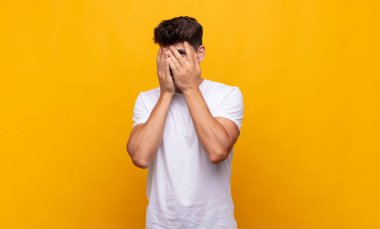 Genç yakışıklı adam elleriyle yüzünü kapatıyor, parmaklarının arasından şaşkın bir ifadeyle yan tarafa bakıyor.