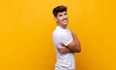 Genç yakışıklı adam neşeyle gülümsüyor, mutlu, tatmin olmuş ve rahatlamış hissediyor, kollarını kavuşturup yan tarafa bakıyor.