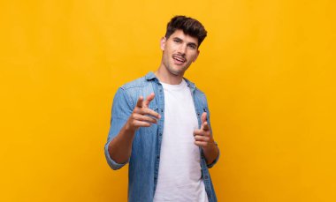 Olumlu, başarılı, mutlu bir tavırla gülümseyen genç yakışıklı adam kamerayı işaret ediyor, elleriyle silah işareti yapıyor.