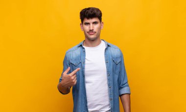 Genç yakışıklı, gururlu, kendinden emin ve mutlu görünüyor, gülümsüyor ve kendini işaret ediyor ya da bir numara işareti yapıyor.