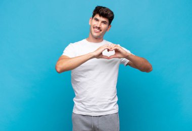 Genç yakışıklı adam gülümsüyor ve mutlu, sevimli, romantik ve aşık, iki eliyle kalbi şekillendiriyor.