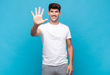 Genç yakışıklı adam gülümsüyor ve arkadaş canlısı görünüyor, 5 ya da 5 numarayı gösteriyor, eli önde, geriye doğru sayıyor.