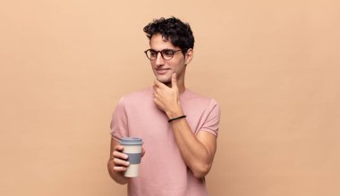 Elinde kahveyle gülümseyen genç yakışıklı bir adam. Gülümseyen, kendine güveni tam, eli çenesinde, merak eden ve yan tarafa bakan bir adam.