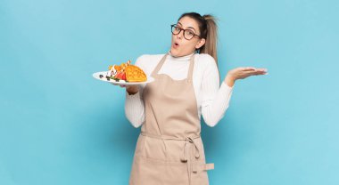 Genç sarışın kadın şaşırmış ve şoka uğramış görünüyor. Çenesi düşmüş ve eli açık bir objeyi tutuyor.