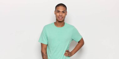 Genç siyah adam mutlu bir şekilde gülümsüyor, eli kalçasında ve kendinden emin, olumlu, gururlu ve arkadaş canlısı.