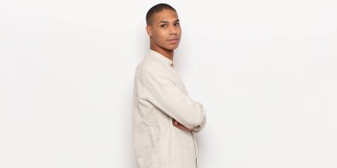 Genç siyah adam kameraya çapraz kollarla gülümsüyor ve mutlu, kendinden emin, tatmin olmuş bir ifade, yan görüş.