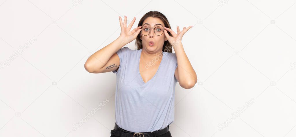 young blonde woman feeling shocked, amazed and surprised, holding glasses with astonished, disbelieving look