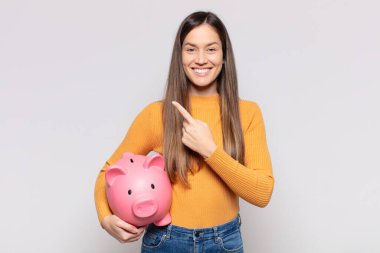 Güzel bir kadın neşeyle gülümsüyor, mutlu hissediyor ve kenarı ve yukarıyı işaret ediyor, fotokopi uzayındaki nesneleri gösteriyor.