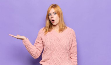 Kızıl saçlı, güzel bir kadın şaşırmış ve şok olmuş görünüyor, çenesi düşmüş, eli açık bir objeyi tutuyor.