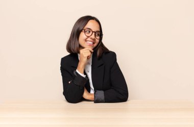 Mutlu, kendinden emin bir ifadeyle gülümsüyor, elleri çenesinde, merak ediyor ve yan tarafa bakıyor.