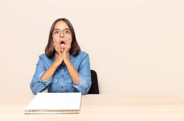 Genç kadın şok olmuş ve korkmuş hissediyor, ağzı açık ve elleri yanaklarında korkmuş görünüyor.