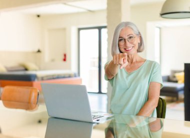 Orta yaşlı bir kadın kameraya bakıyor, kendinden emin, arkadaş canlısı bir gülümsemeyle, seni seçiyor.