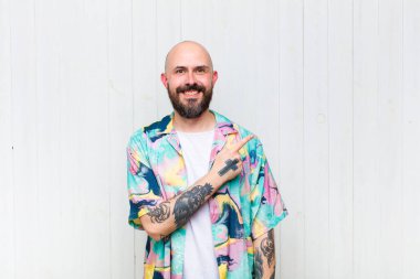 Kel adam neşeyle gülümsüyor, mutlu hissediyor ve kenarı ve yukarıyı işaret ediyor, fotokopi alanında nesneler gösteriyor.