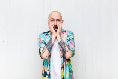 Kel adam şok olmuş ve korkmuş hissediyor, ağzı açık ve elleri yanaklarında korkmuş görünüyor.