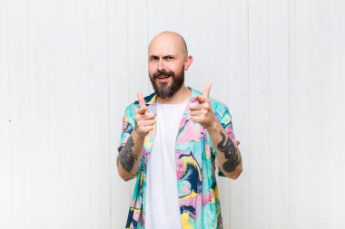 Olumlu, başarılı, mutlu bir tavırla gülümseyen kel bir adam kamerayı işaret ediyor, elleriyle silah işareti yapıyor.