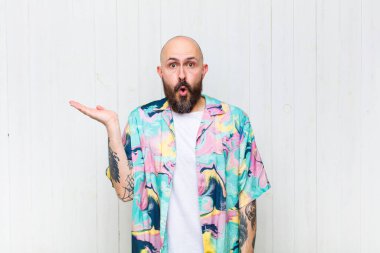 Kel adam şaşırmış ve şoka uğramış görünüyor, çenesi düşmüş ve eli açık bir nesneyi tutuyor.