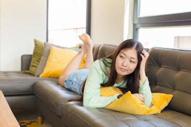 Genç Asyalı kadın şaşkın ve kafası karışmış hissediyor, başını kaşıyor ve yan tarafa bakıyor.