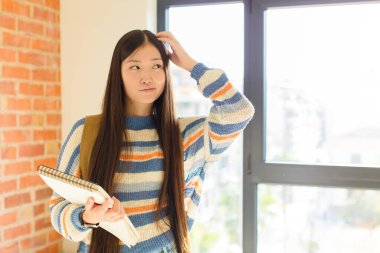 Genç Asyalı kadın şaşkın ve kafası karışmış hissediyor, başını kaşıyor ve yan tarafa bakıyor.