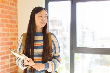 Asyalı genç bir kadın mutlu, kendinden emin bir ifadeyle gülümsüyor, çenesinde elleri, merak ediyor ve yan tarafa bakıyor.