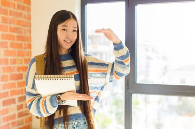 Genç Asyalı kadın gülümsüyor, mutlu, pozitif ve tatmin olmuş hissediyor, fotokopi alanında nesne veya konsept tutuyor veya gösteriyor
