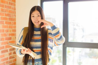 Genç Asyalı kadın gülümsüyor ve mutlu, sevimli, romantik ve aşık, iki eliyle kalbi şekillendiriyor.